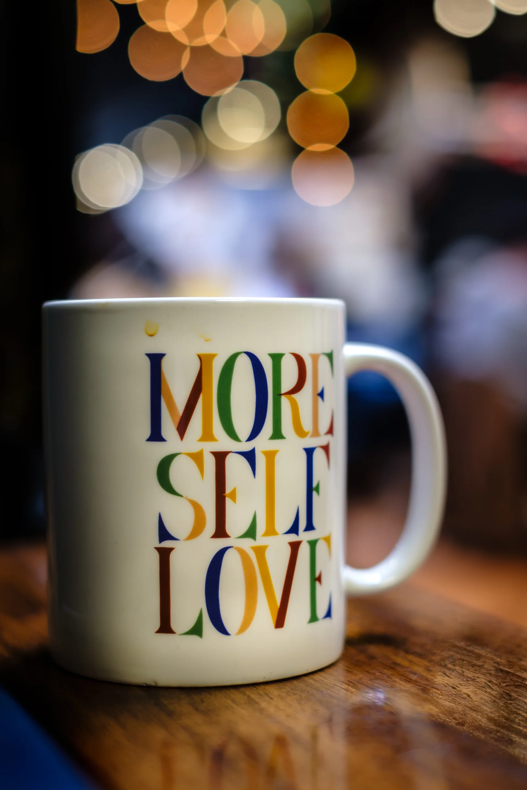 Brand & location — coffee mug reading "More Self Love", warm café bokeh
